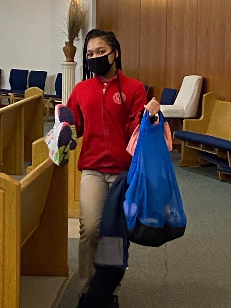 student carrying a blue bag and holding shoes behind church pews